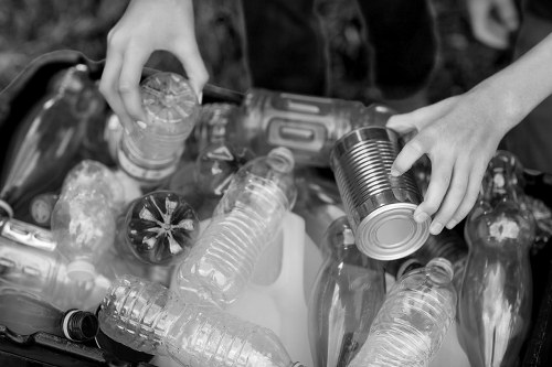 A person’s hands sorting through a collection of various used plastic bottles and cans, including clear soda bottles, a metal can, and other recyclable containers, placed in a container or bin outdoors. The bottles are of different shapes and sizes, some with labels or caps, and display a mix of transparent plastic and metallic finishes. The background is blurred, suggesting an environment dedicated to waste collection or recycling activities, possibly on a pavement or driveway. The scene is captured in black and white, emphasizing the textures of the plastic and metal surfaces. This image illustrates proper handling of recyclable rubbish, consistent with waste removal services offered by companies like Office Clearance Feltham. The focus on discarded bottles and cans highlights the importance of waste segregation in local rubbish clearance efforts, especially in the Feltham area, which can be part of a professional waste management process.