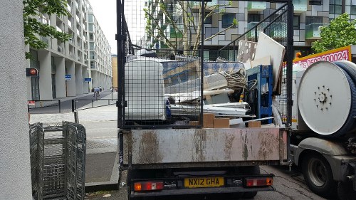 A flatbed refuse collection vehicle parked on a city street with various types of rubbish loaded onto its cargo area, including broken furniture, white panels, metal framing, and other miscellaneous waste materials. The truck's surface appears rusted and weathered, with some visible dirt and grime. In the background, there are modern multi-storey residential buildings with numerous windows and balconies, along with a pedestrian crossing signal visible at the intersection. The scene suggests a rubbish removal operation, possibly for a property clearance service in Feltham or nearby in southwest London, with the vehicle positioned near a corner of the road and adjacent to a sidewalk. The environment looks urban and busy, with the waste materials arranged in a manner typical of commercial or residential clearance activities, handled by companies such as Office Clearance Feltham.