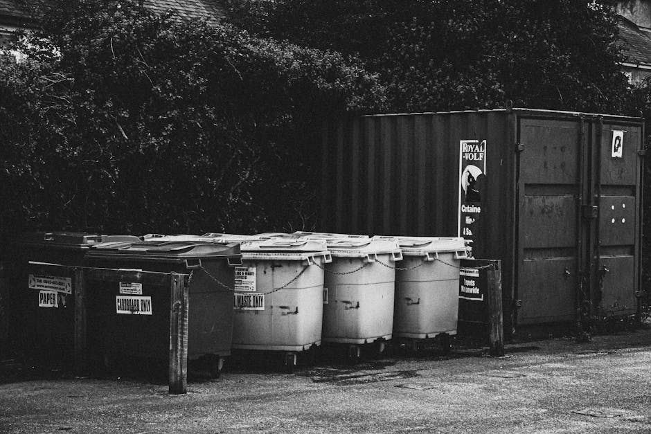 A large, blue plastic wheelie bin with a closed lid is positioned on a concrete pavement next to the side of a white delivery truck. The bin appears to have a slightly textured surface and is stained