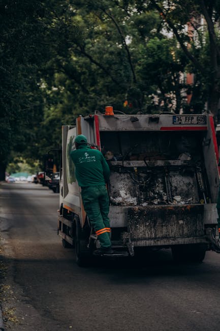 A worker dressed in a green uniform with reflective orange stripes is seen from the back, standing beside a waste collection vehicle on a quiet street lined with trees. The vehicle, which appears to b