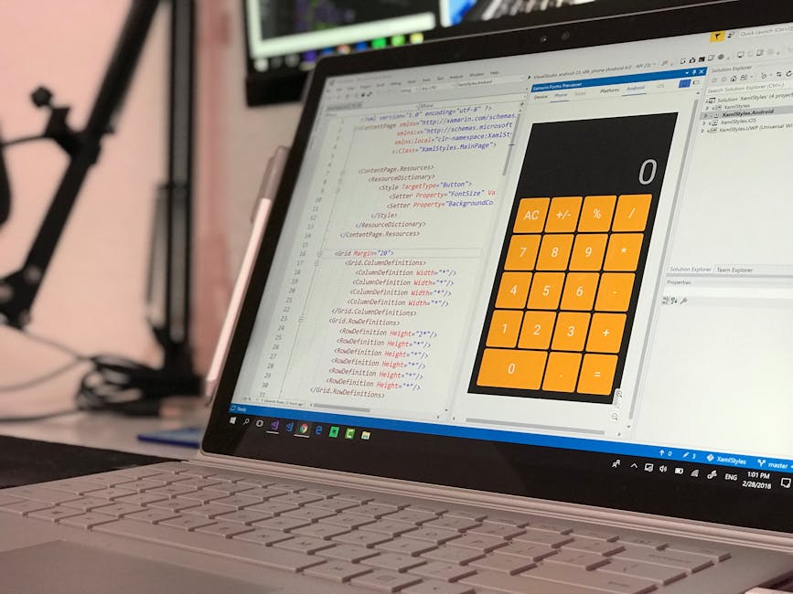 A person in a tan long-sleeved shirt is working on a laptop at a cluttered desk, with their right hand holding a stylus and their left hand pointing at the screen. The laptop displays lines of code in